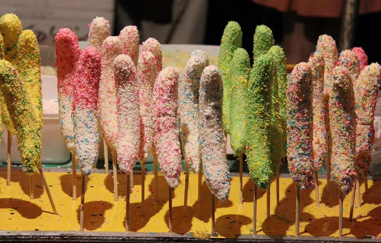 Rows of bananas on sticks, coated in white, pink and green chocolate and covered with sprinkles.