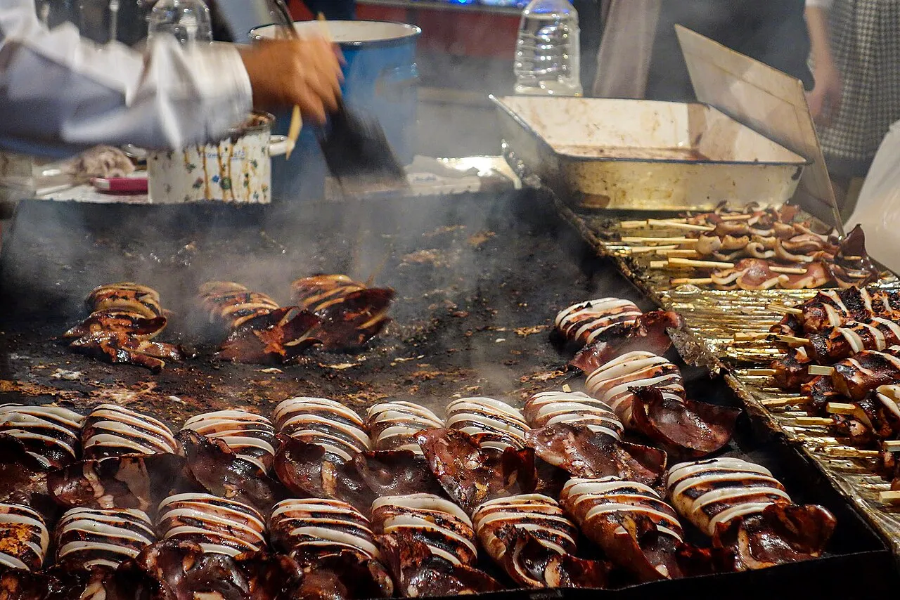 Whole squid and squid skewers grilling on a smoking iron hotplate at a food stall.