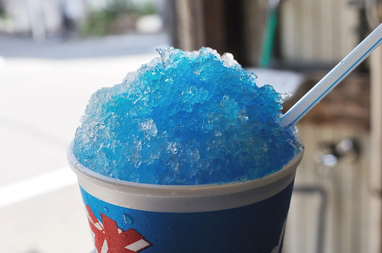 A paper cup heaped with shaved ice soaked in bright blue syrup, with a straw spoon.