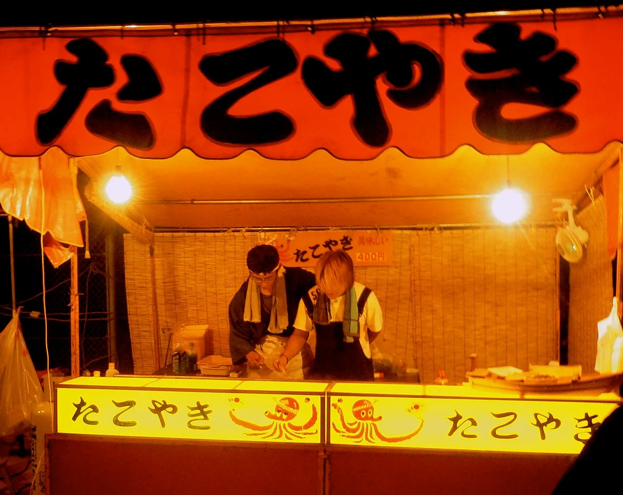 A brightly lit takoyaki stall at night, with an octopus drawn on the glowing counter and two cooks at work behind it.