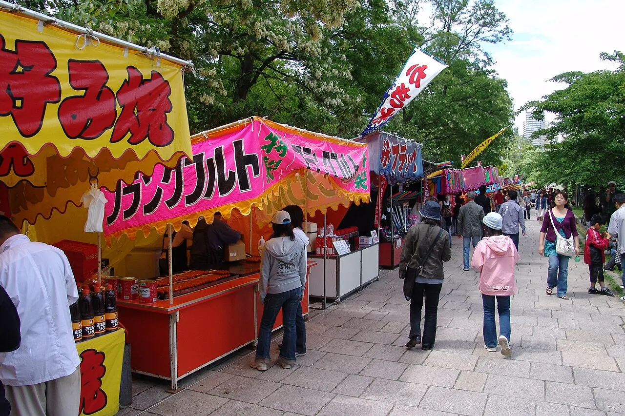 A row of colourful food stalls with big painted banners along a tree-lined park path, with people walking past.