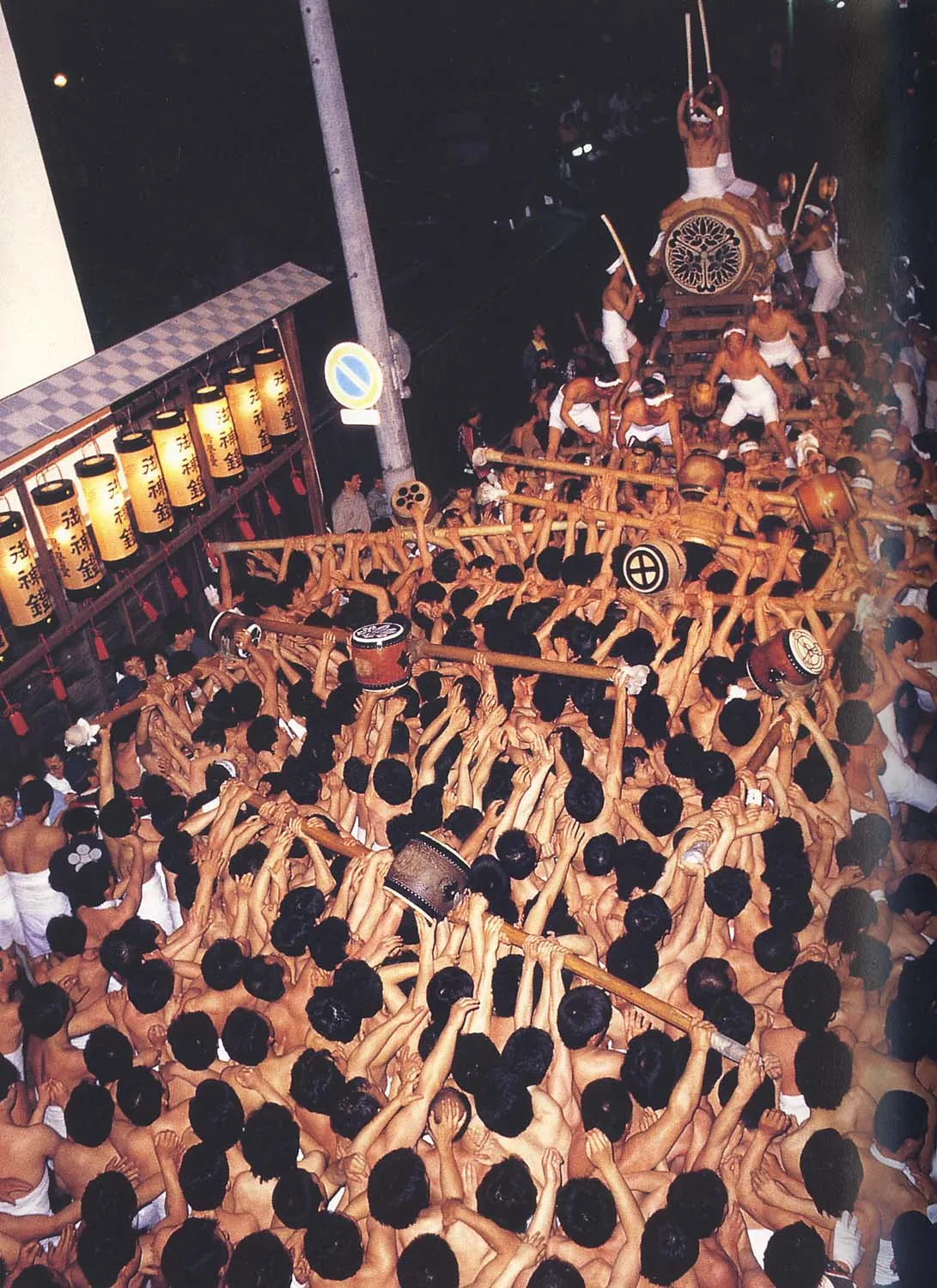 A night crowd of men in white wrappings pressing around the frame of the Okoshi Daiko drum, seen from above.