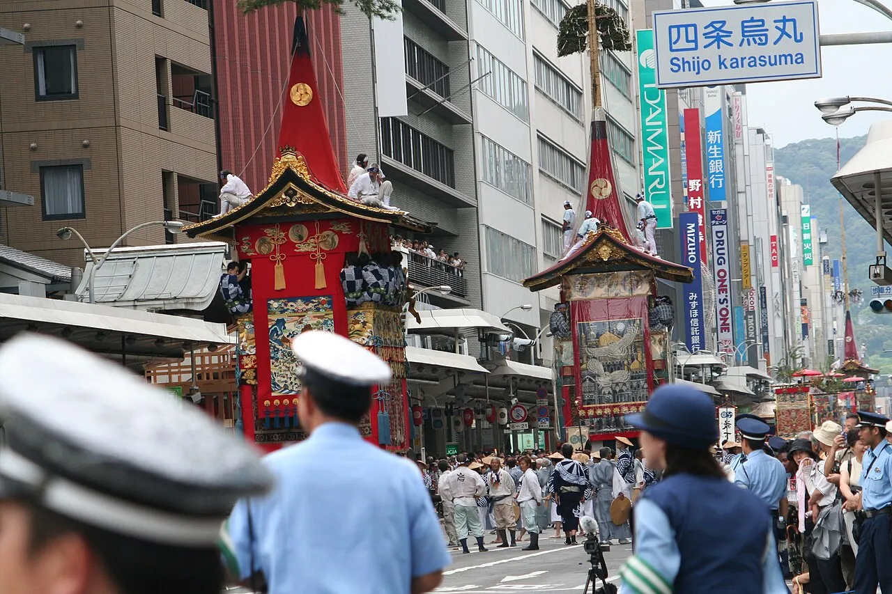 Two tall red hoko floats move along Shijō Street at the Shijō Karasuma intersection, with crowds and police lining the road.