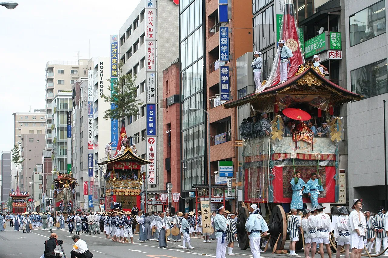 The Iwatoyama float, with a pointed roof and red and gold hangings, rolls down a Kyoto street lined with office buildings.
