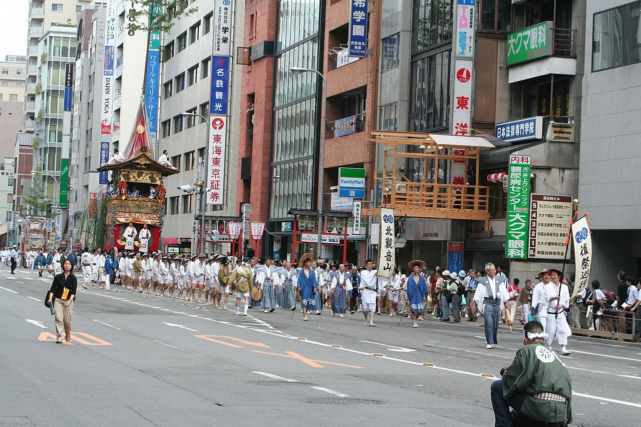 Men in white and blue robes and straw hats walk along a wide Kyoto street ahead of a float, carrying a banner.