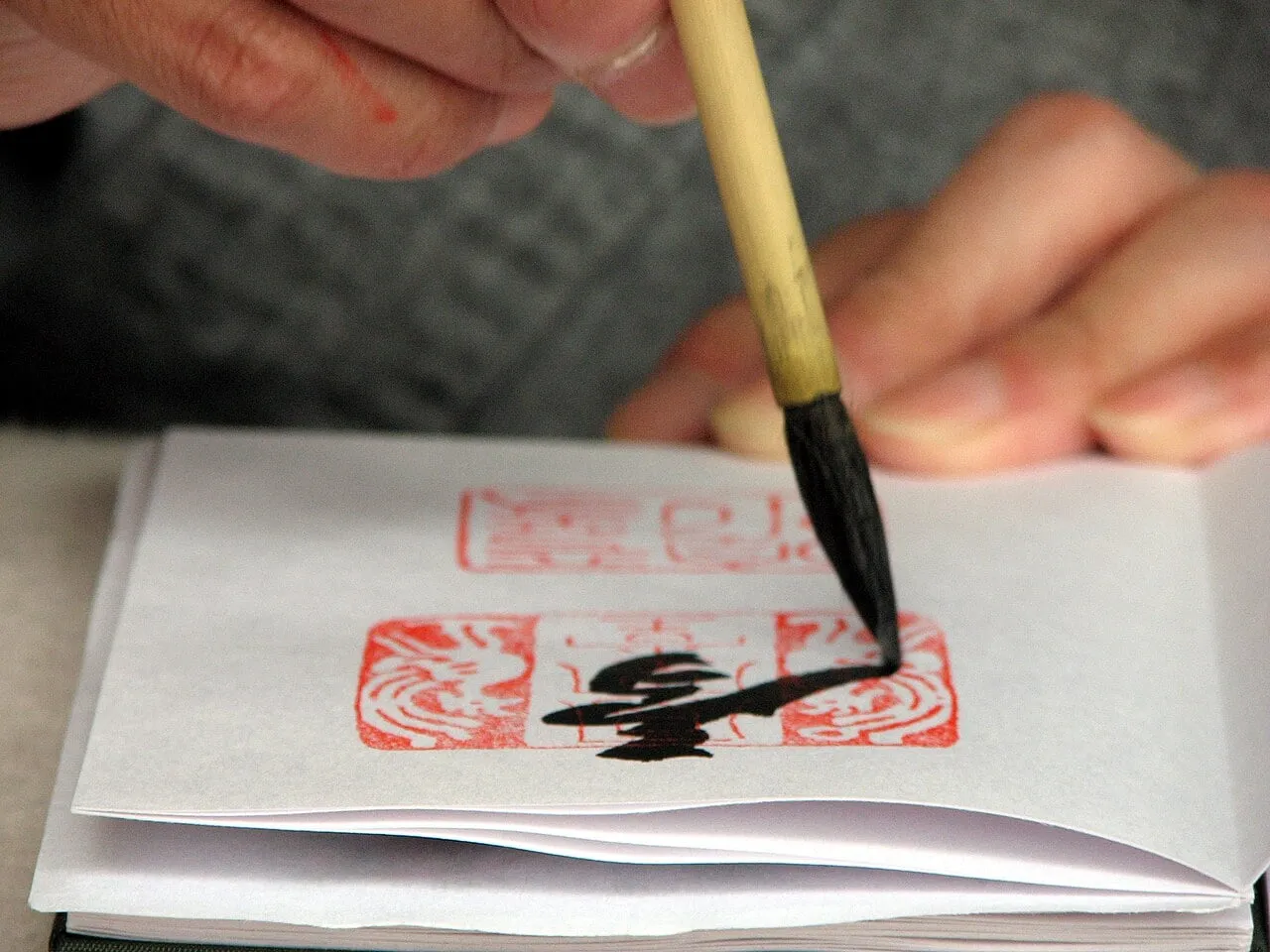 A hand holding a brush writes black characters over red seals on a page of a goshuin book.