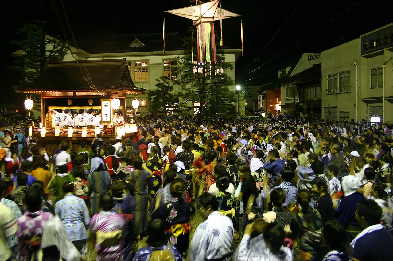 A packed crowd in yukata dances in a square at night around a lantern-hung music float, under a tall lantern pole.