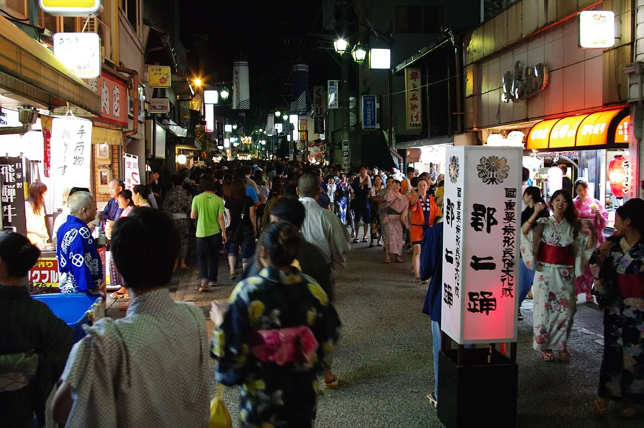 People in yukata dance down a narrow, lantern-lit street of old shops late at night.