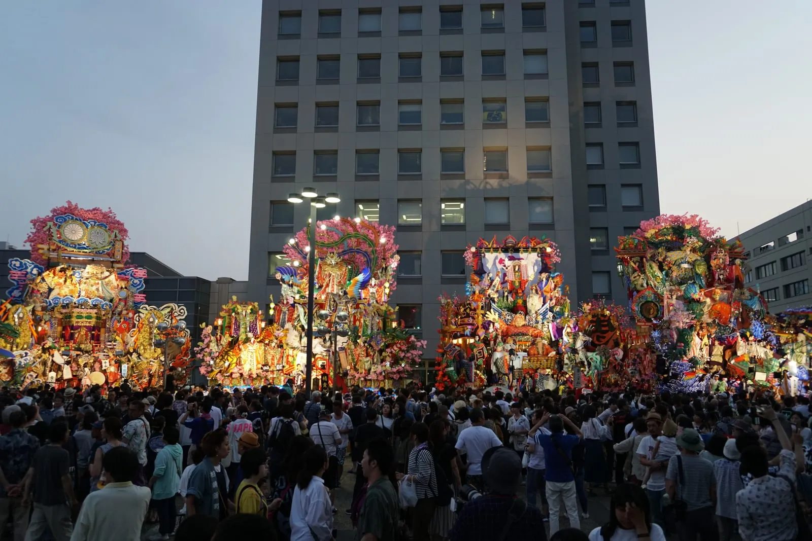A row of tall, colourful floats decorated with figures and flowers stands in front of an office building as crowds gather.