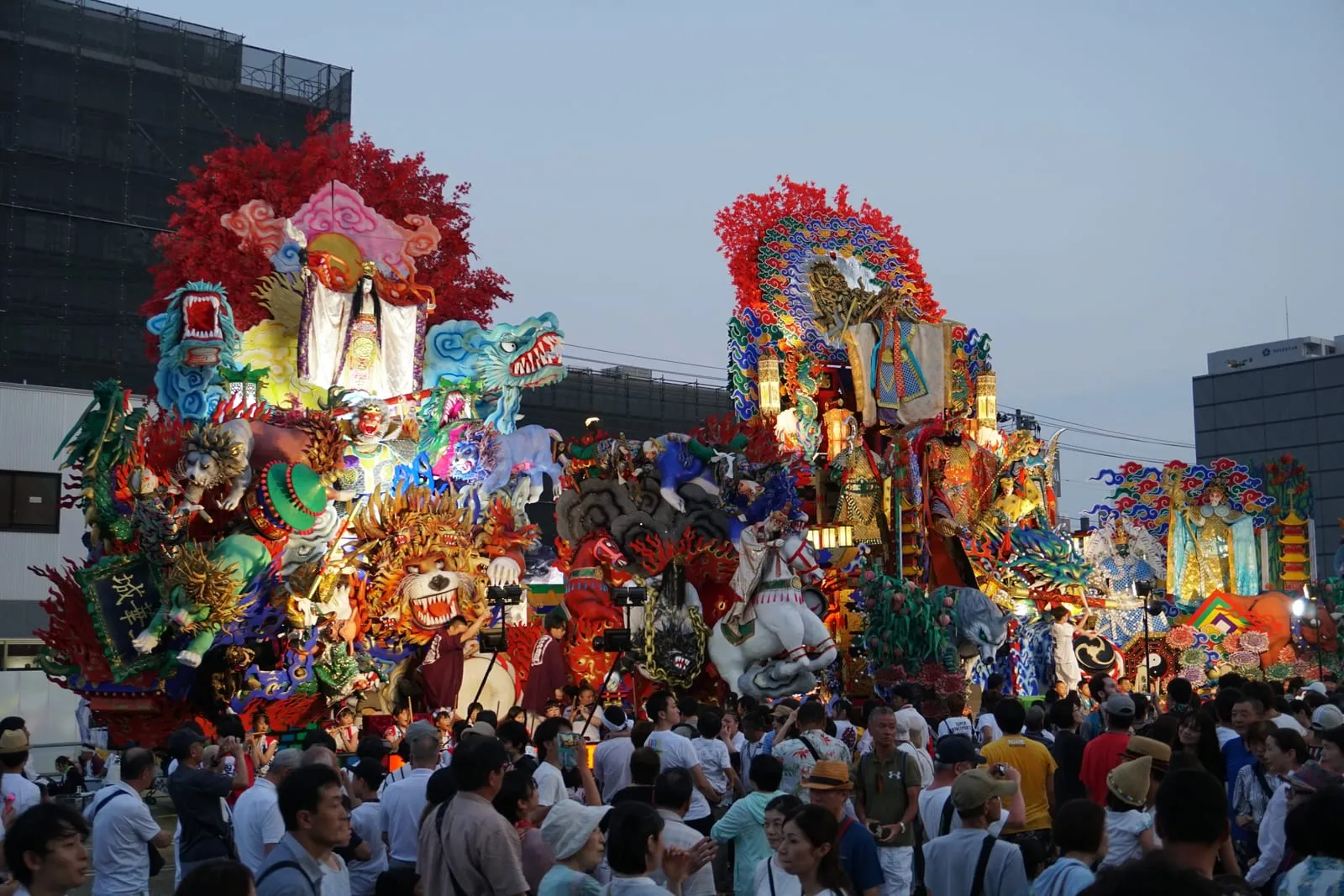 Close view of two towering floats covered in sculpted figures, dragons and red blossoms above a crowd.