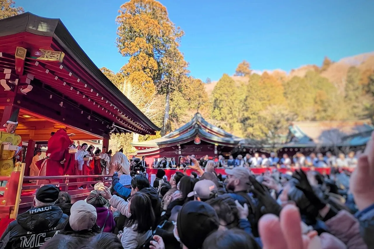 A crowd reaches up with open hands towards people throwing beans from a red shrine hall.