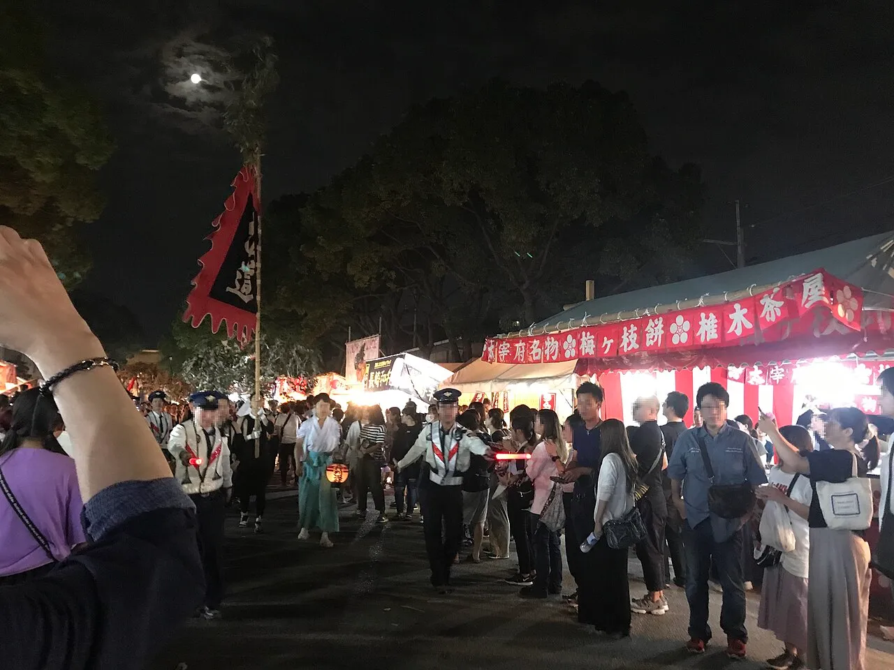 Crowds walk past lit festival stalls along the shrine approach at night, under a full moon.