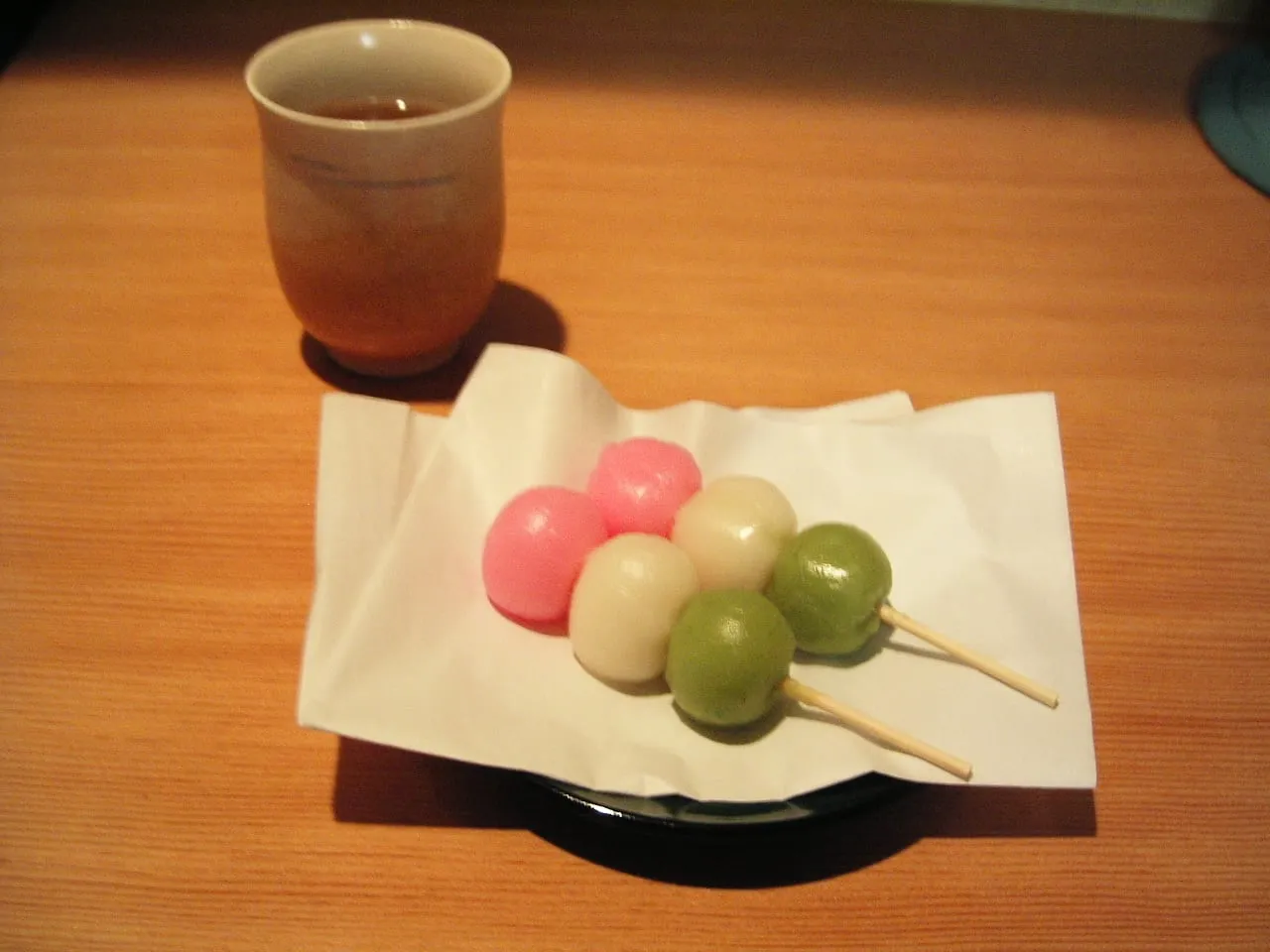 A skewer of three rice dumplings, pink, white and green, on paper next to a cup of tea.