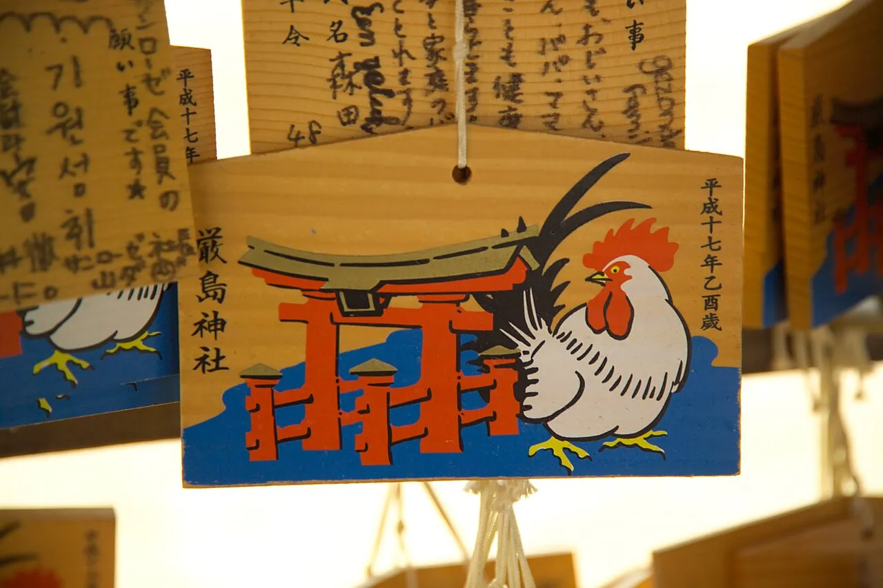 A wooden ema plaque painted with a red torii gate in the sea and a rooster, hanging among plaques written with wishes.