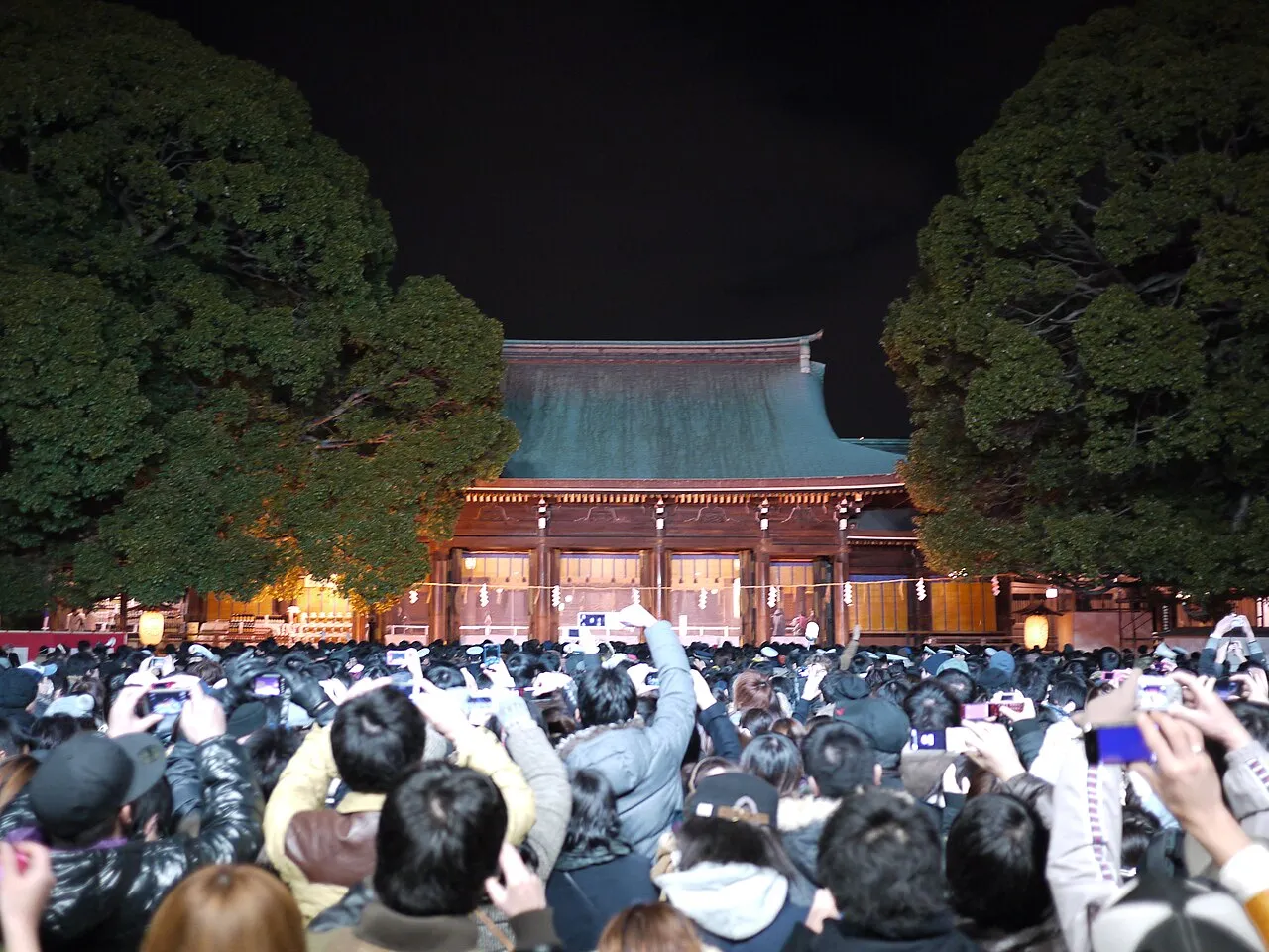 A dense crowd at night, many holding up phones, faces the lit main hall of Meiji Jingū between two large trees.