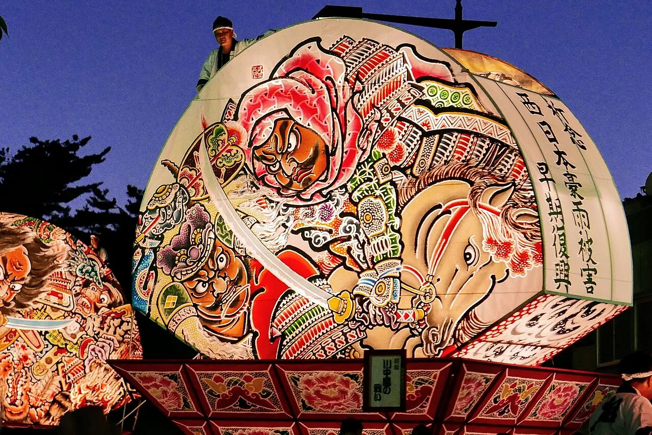 A giant fan-shaped paper float, lit from inside, painted with a fierce warrior on horseback, against an evening sky.