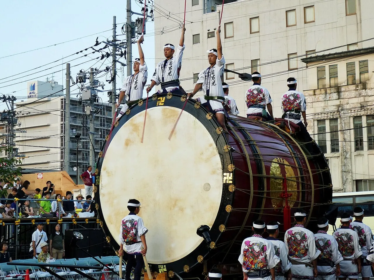 Drummers stand on top of a huge drum on a float, raising their sticks in a city street.