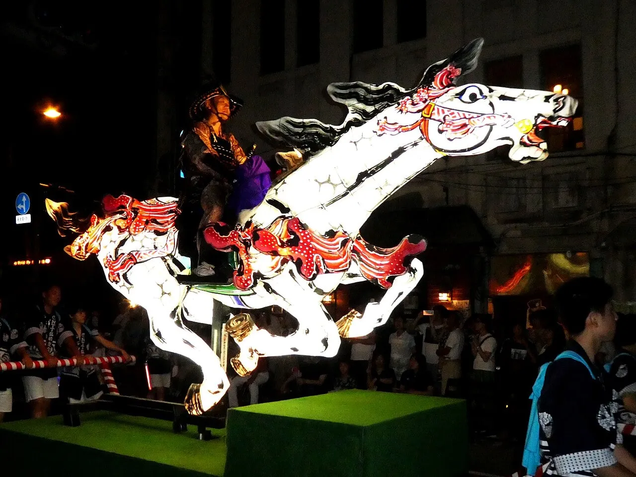 A glowing paper float in the shape of a white horse with a warrior rider, at night.