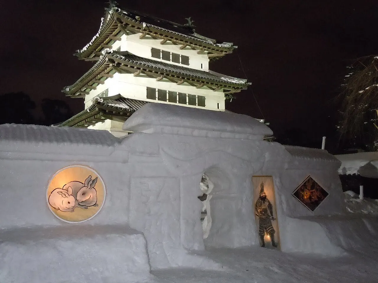 A wall of carved snow with round and diamond-shaped windows, lit at night below a model of a castle tower.