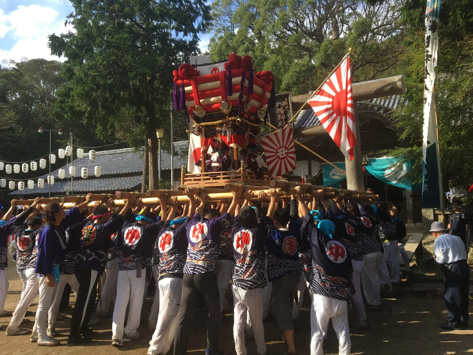 Men in matching coats lift a red and gold chosa float on their shoulders in front of a shrine, with a large flag behind.