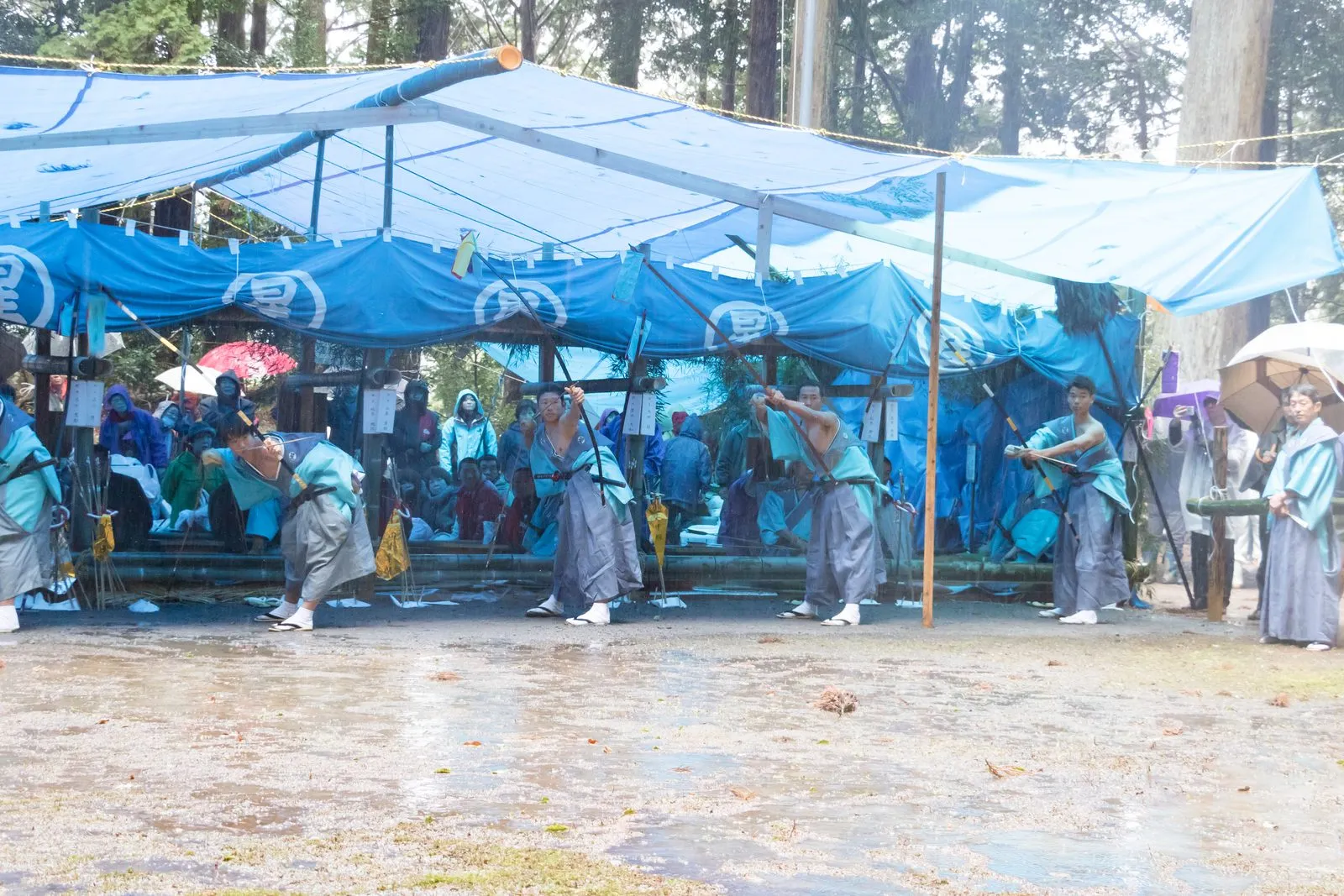 Archers in traditional dress stand in a rain-soaked shrine ground under blue tarpaulins.