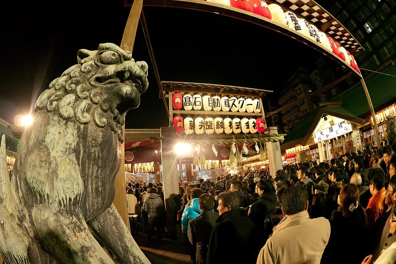 Crowds fill Imamiya Ebisu Shrine at night under rows of paper lanterns, with a stone guardian lion in the foreground.