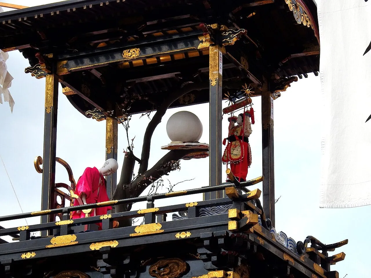 A puppet in red robes stands on the top tier of a black-lacquered float with gold fittings.