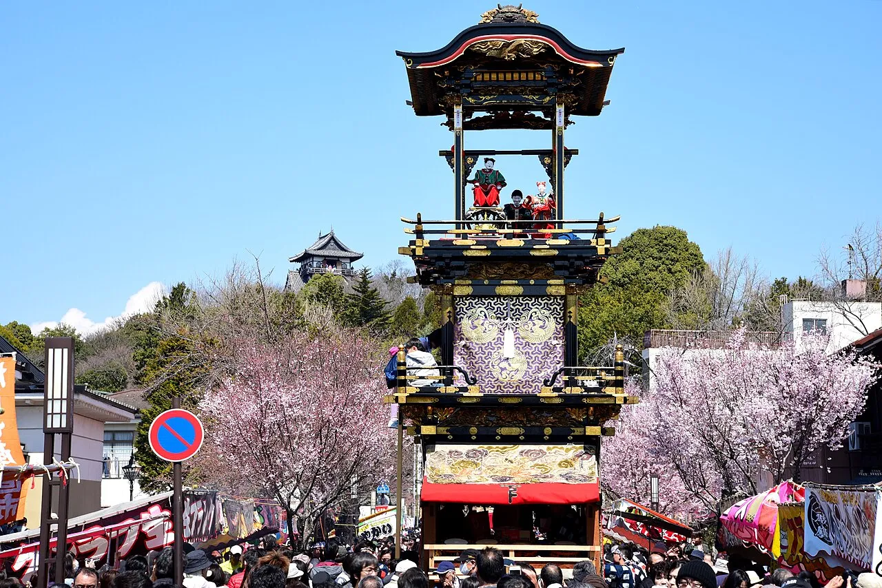 A tall three-tiered wooden float with a red curtain stands among cherry blossoms, with Inuyama Castle on the hill behind.