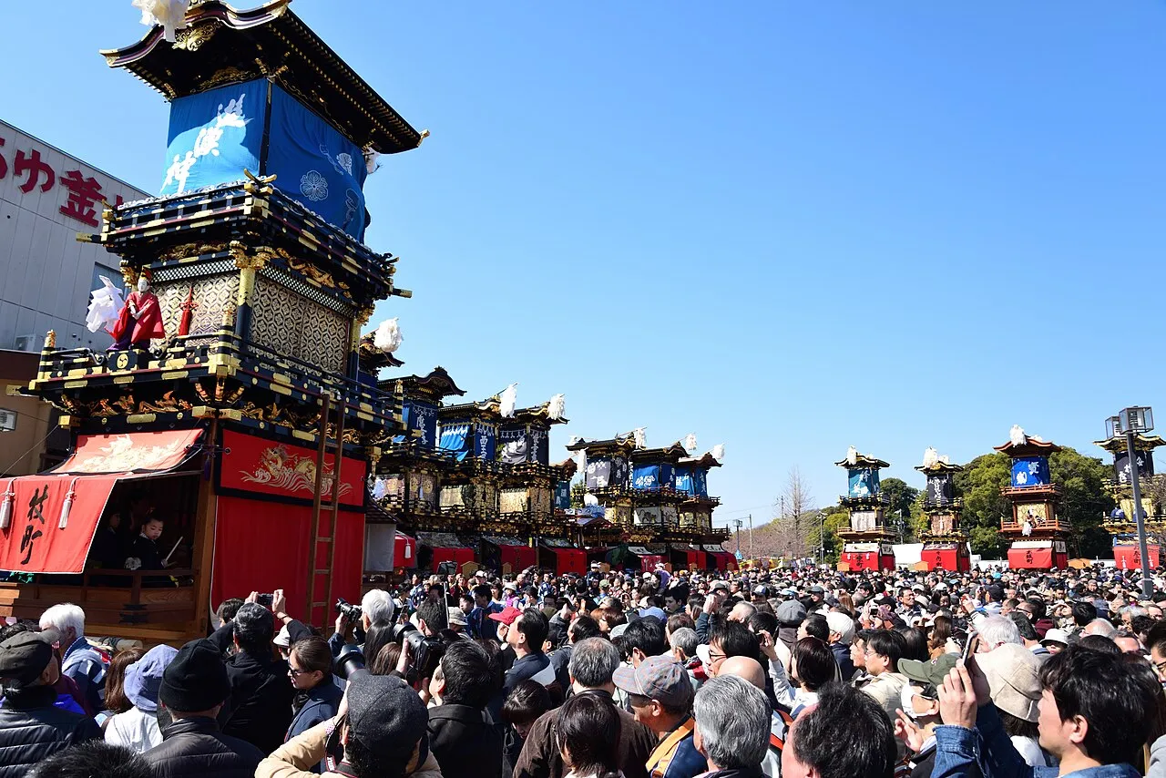 A row of ornate floats lines a street packed with spectators under a clear blue sky.