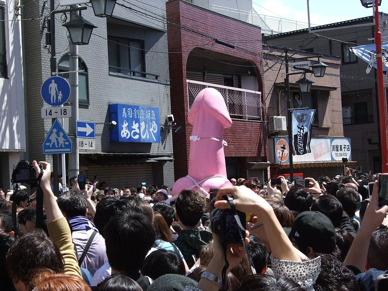 A large pink mikoshi rises above a dense crowd holding up cameras in a narrow street.
