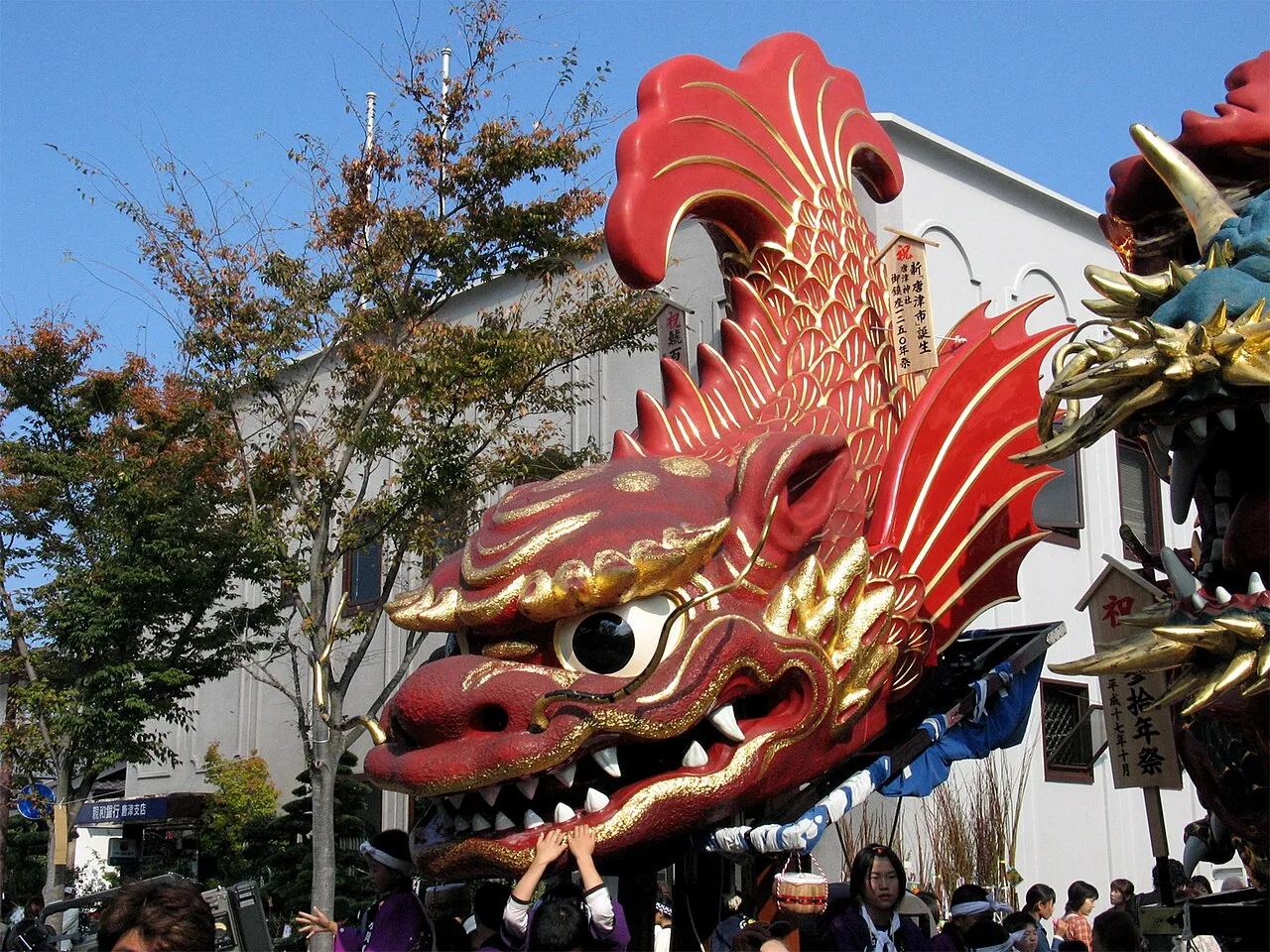 A huge red and gold float shaped like a mythical fish with bared teeth is pulled down a street past onlookers.