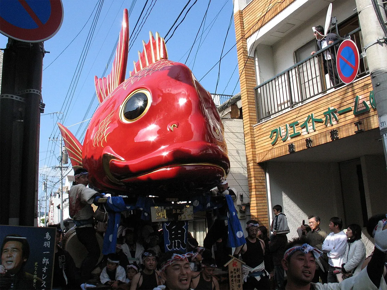 A giant red sea bream float with a staring eye rises above a crowd in a narrow street.