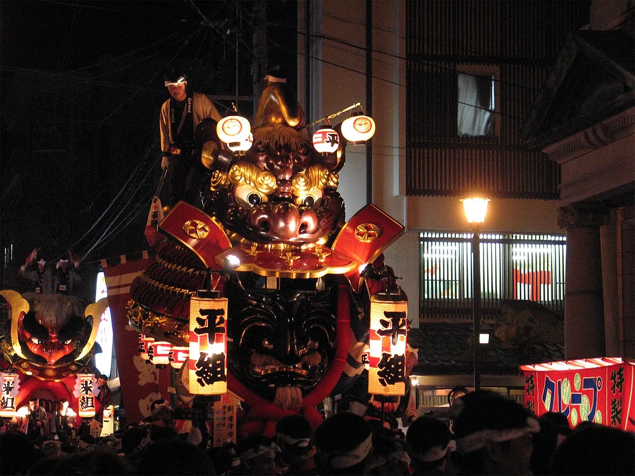 A giant lacquered samurai helmet float lit up at night, with a red-faced lion float behind.