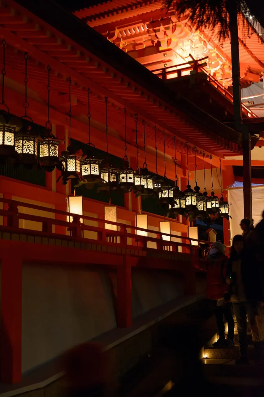 Rows of glowing bronze lanterns hang along a vermilion shrine corridor at night.