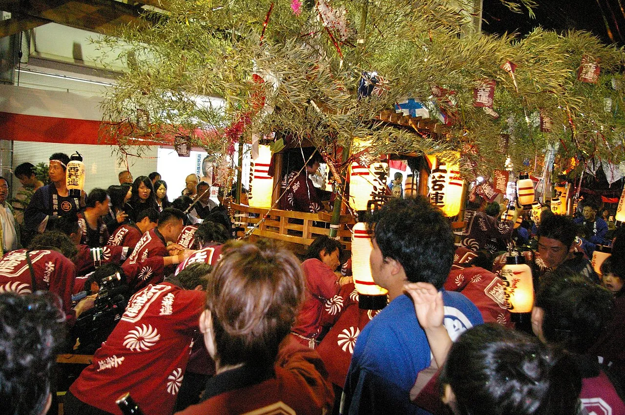 A lantern-lit danjiri float surrounded by men in red festival coats at night.