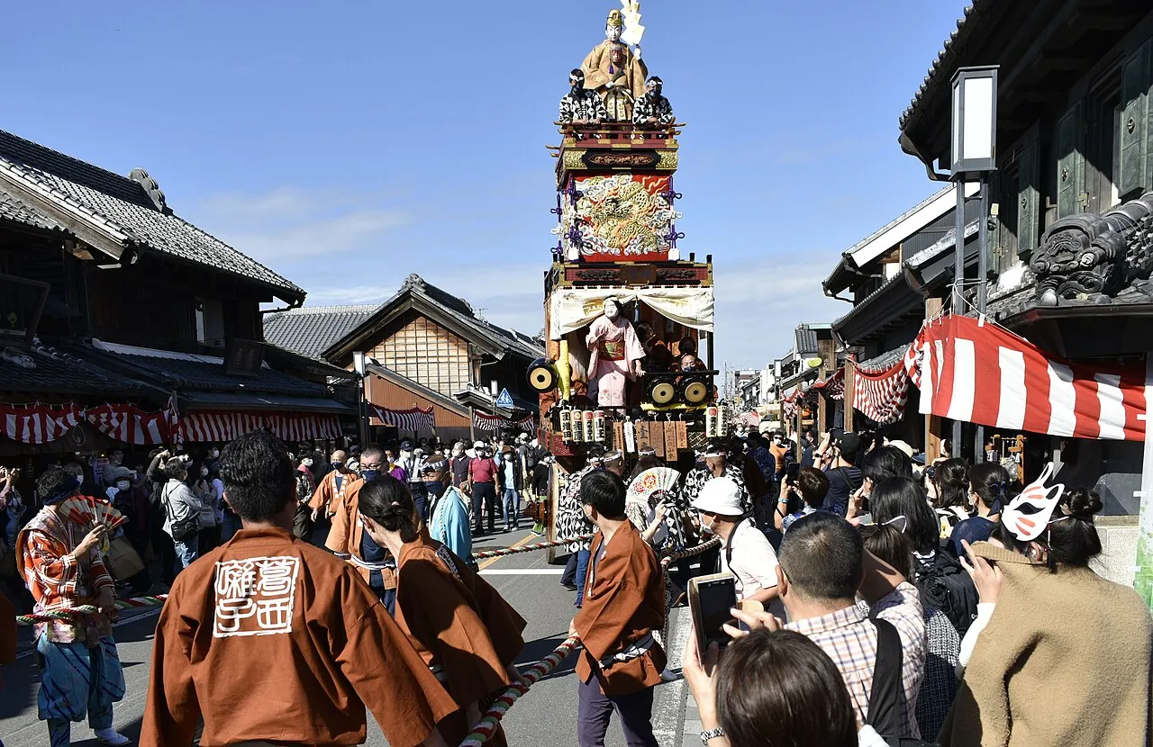 A tall decorated float with a figure on top rolls along a street of old warehouses, surrounded by crowds in festival coats.