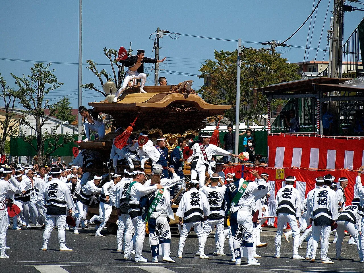 A wooden danjiri cart with a man standing on its roof, pulled by a team in white and black happi coats.