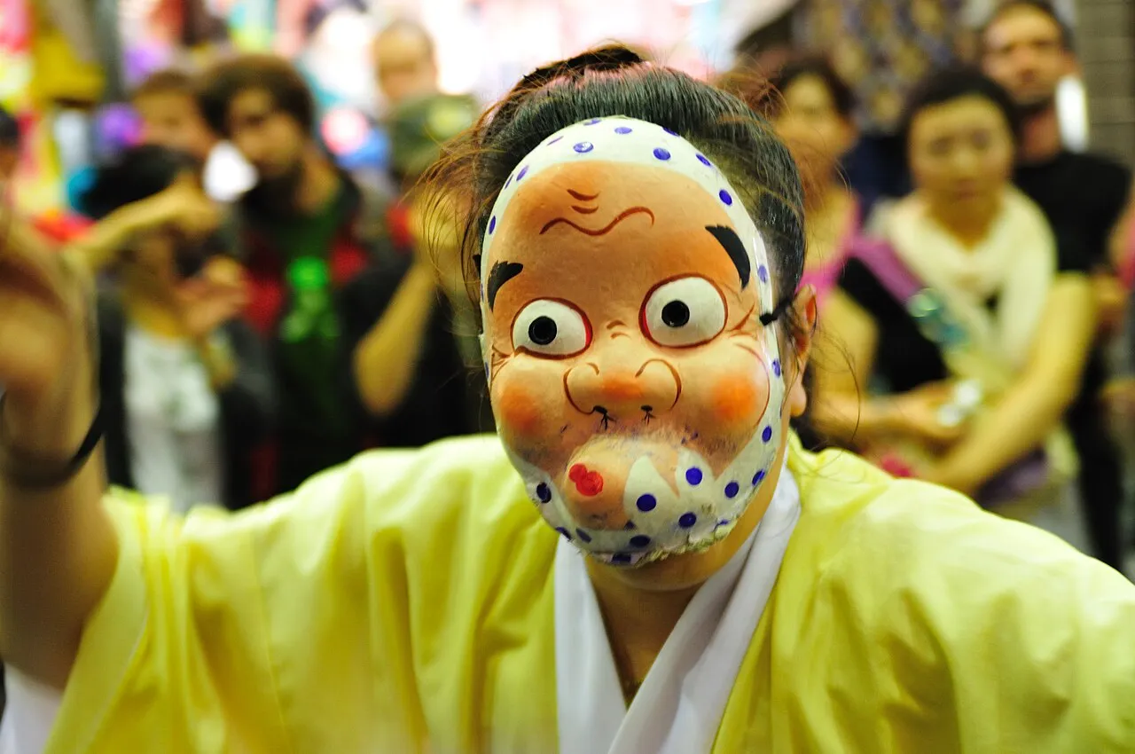 A dancer in a yellow costume and a comic white mask poses in front of a blurred crowd.