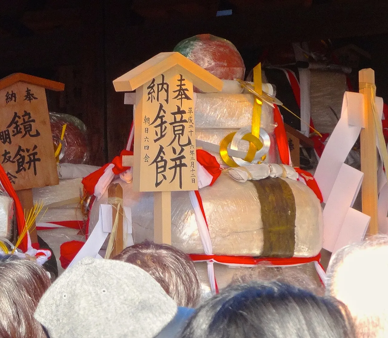 A huge stacked rice cake wrapped in film and tied with red and white cords, with a wooden sign reading 'offering of kagami mochi'.