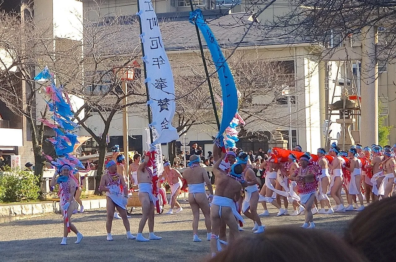 Men in loincloths carry tall bamboo poles hung with paper streamers into the shrine grounds.