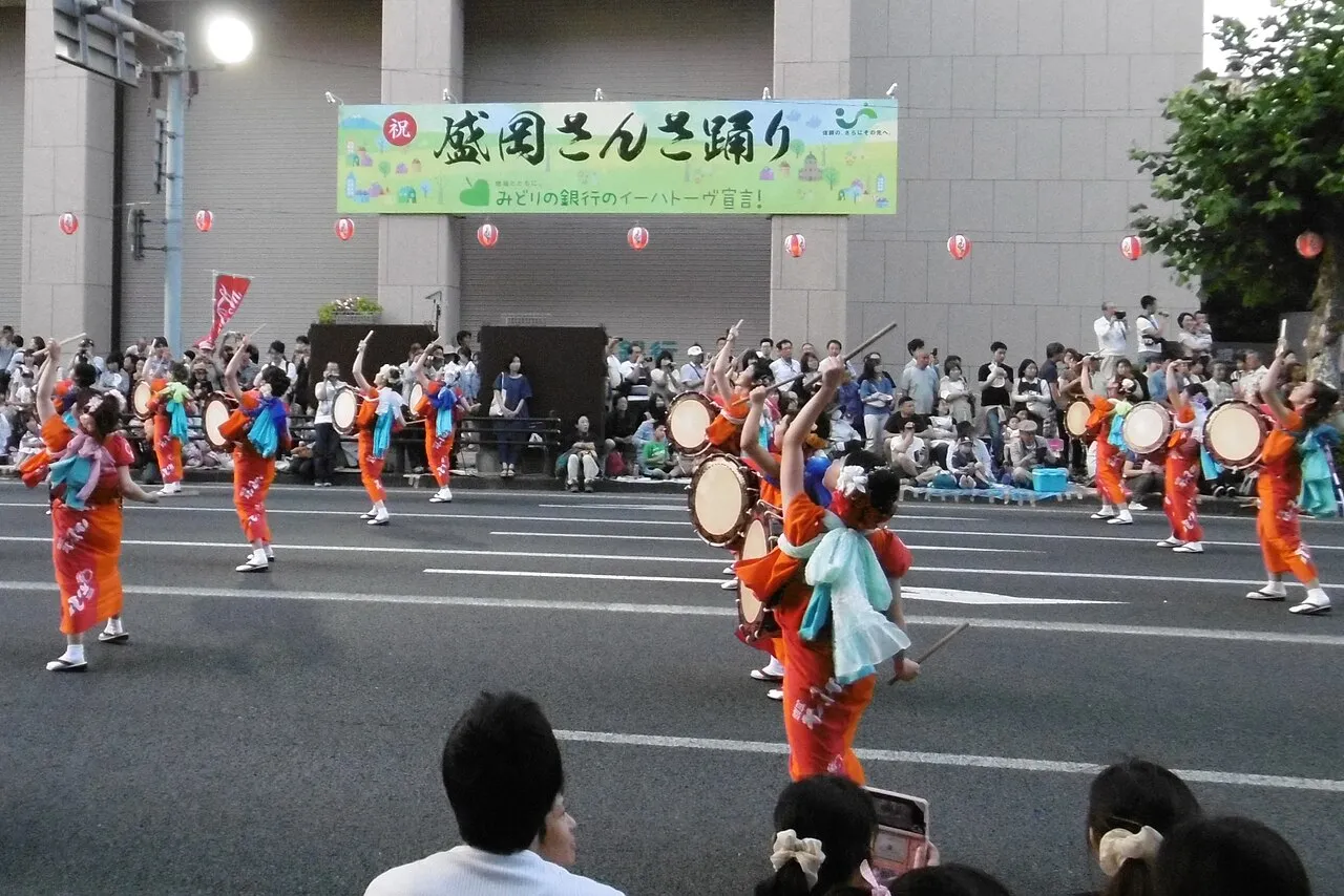 Dancers in red and orange costumes with drums parade along a wide street under a festival banner.