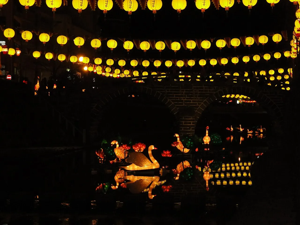 Strings of yellow lanterns glow over a river at night, with lantern figures reflected in the water beside a stone arch bridge.