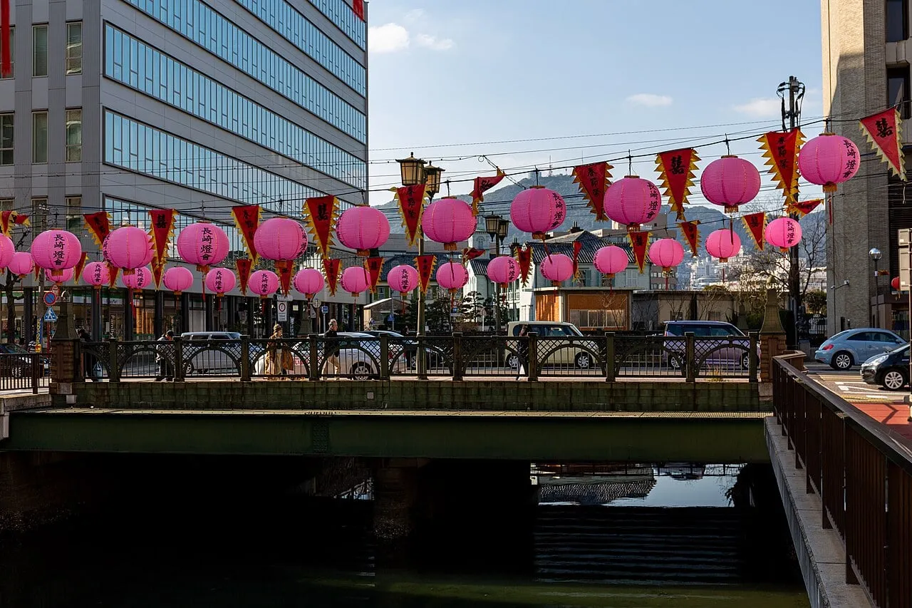 Rows of pink and red Chinese lanterns strung above a small river and bridge in the city.