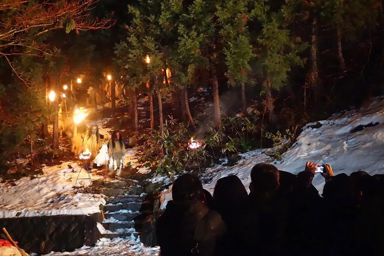 Torches light a line of figures coming down a snowy forest path at night, watched by a crowd.