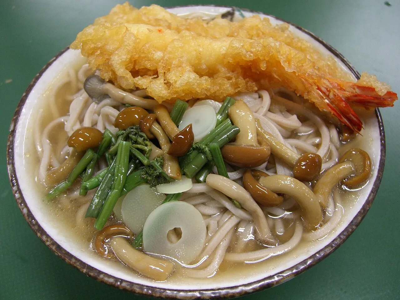 A bowl of buckwheat noodles in broth topped with a large prawn tempura, mushrooms and mountain greens.