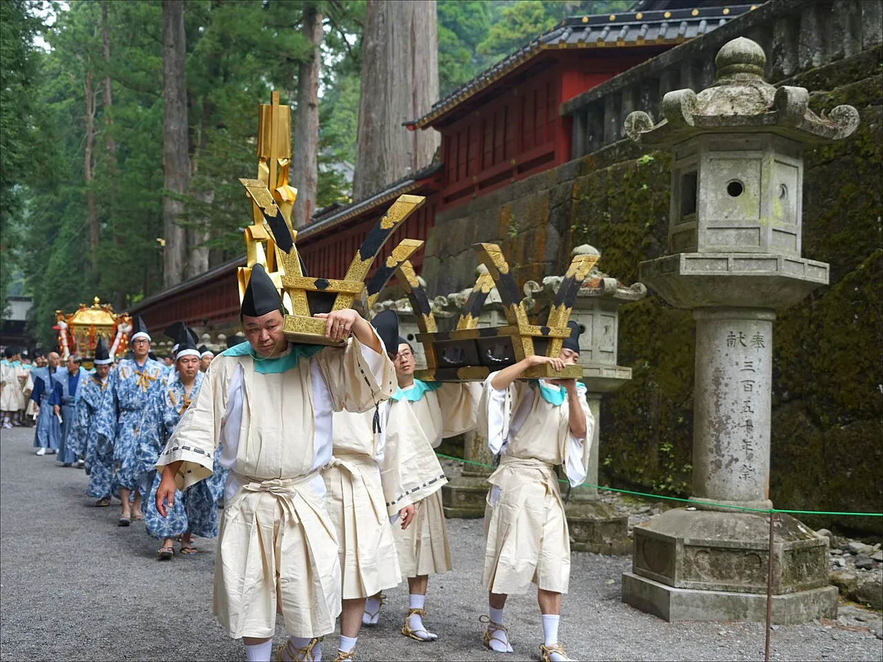 Men in white robes carrying tall golden ceremonial objects in a procession past stone lanterns and a red wall.