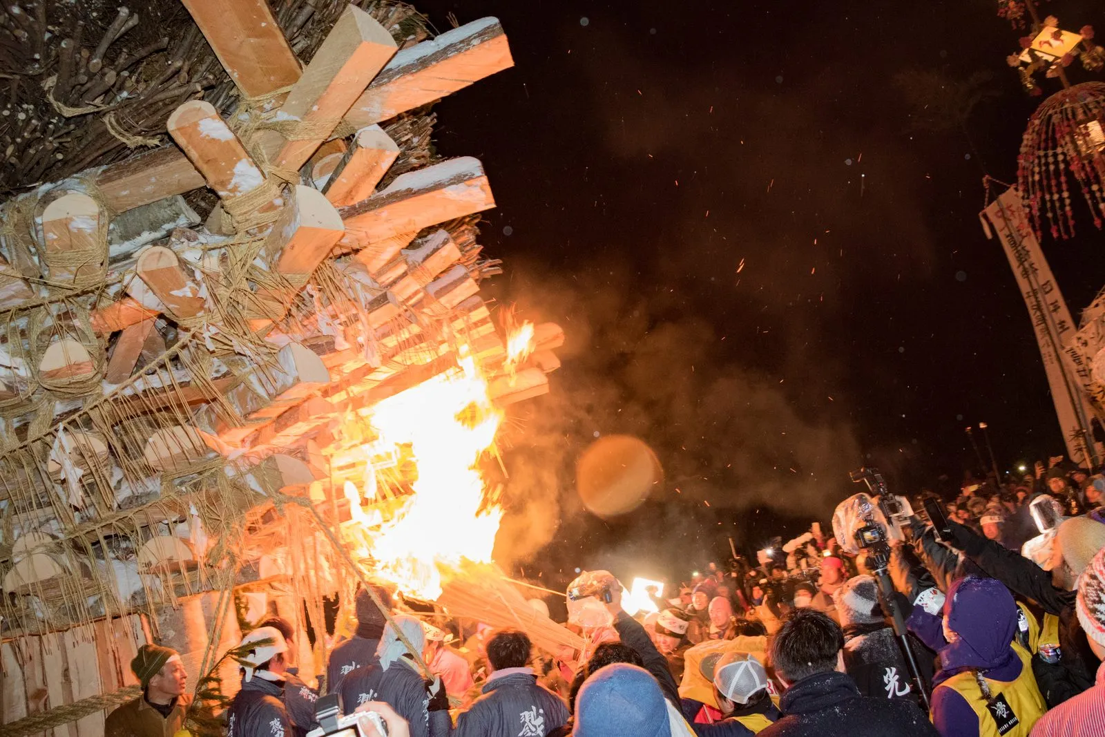 Flames and sparks burst from a wooden shrine structure at night as a crowd presses around it.