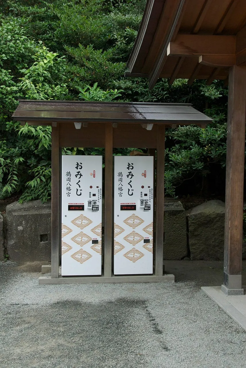Two white omikuji vending machines under a small wooden roof in a shrine garden.