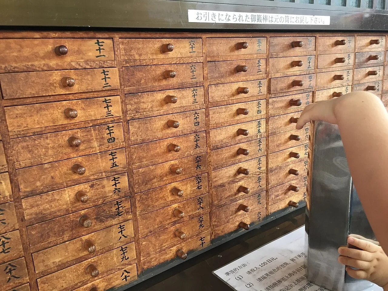 A wall of small numbered wooden drawers; a visitor holds a metal box of fortune sticks.