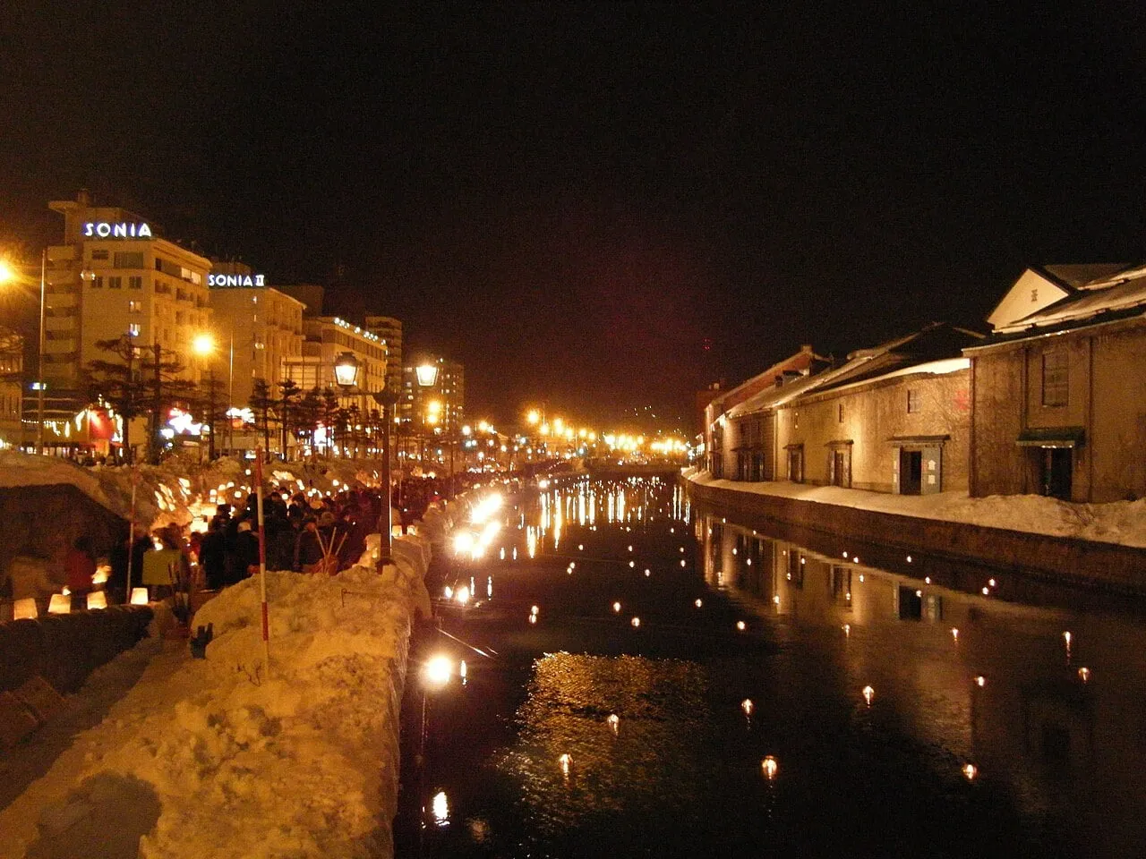 Rows of candles glow in the snow along a canal lined with old stone warehouses at night.